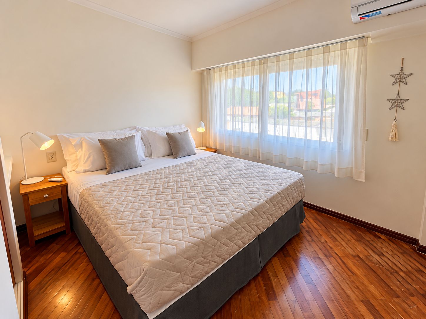 Quarto com cama, mesas de cabeceira, janela com cortinas e ar-condicionado fixado na parede.