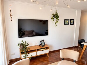 Een woonkamer met een flatscreen televisie aan de muur, een houten consoletafel met lades, twee stoelen en hangende planten.