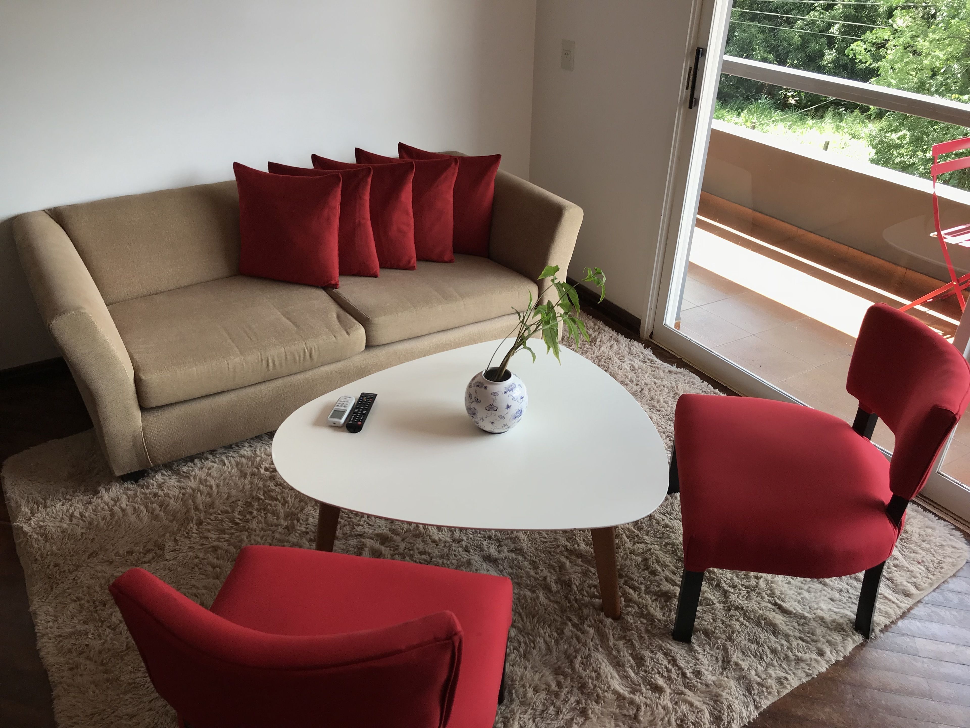 A living room with a beige sofa, red cushions, a white coffee table, a red chair, and a potted plant.