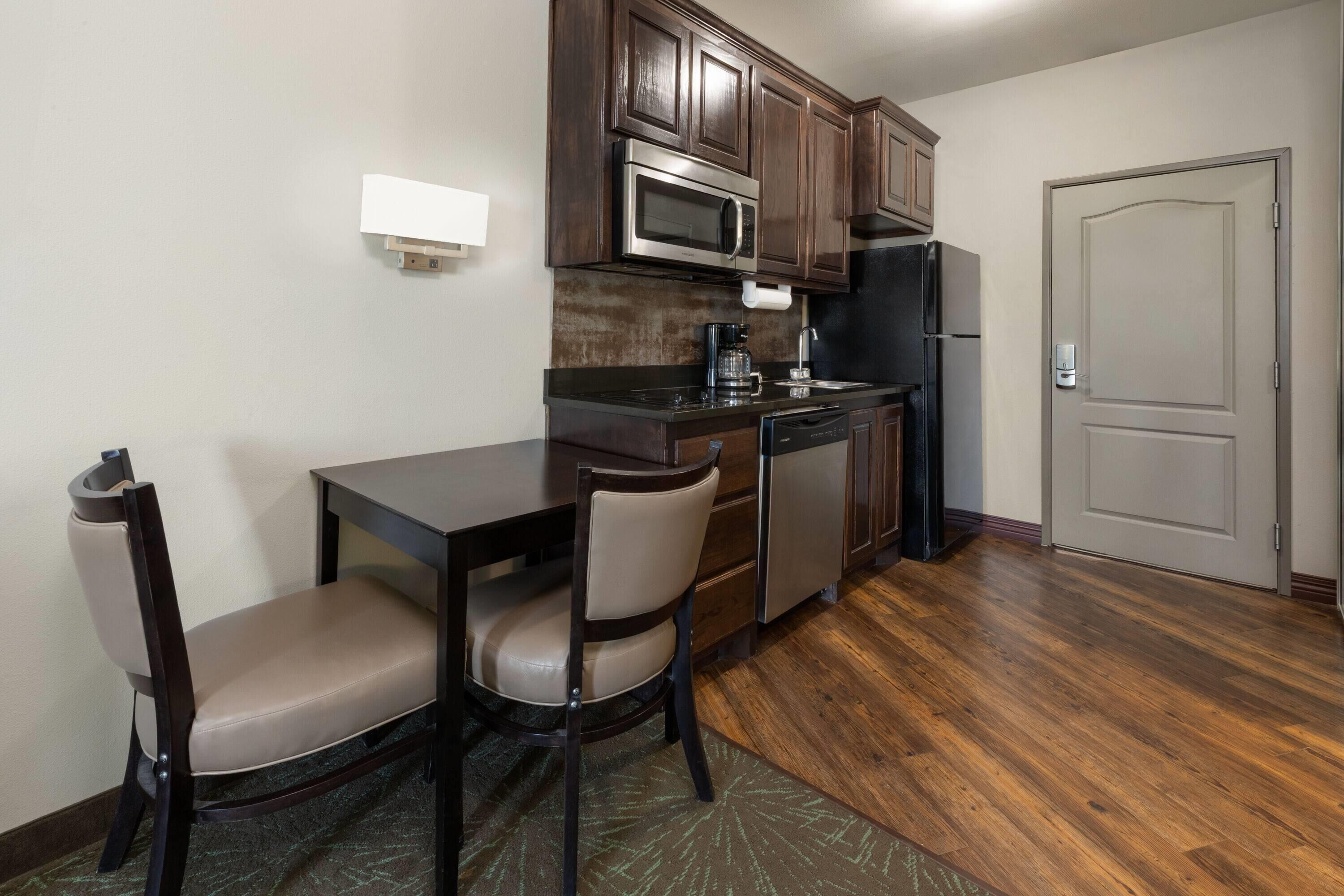 A compact kitchen with dark wooden cabinets, a microwave, a coffee maker, and a refrigerator. There is a dining table with two chairs.
