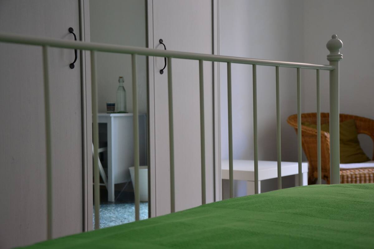 A room with a green bed, white wardrobe, and a wicker chair.