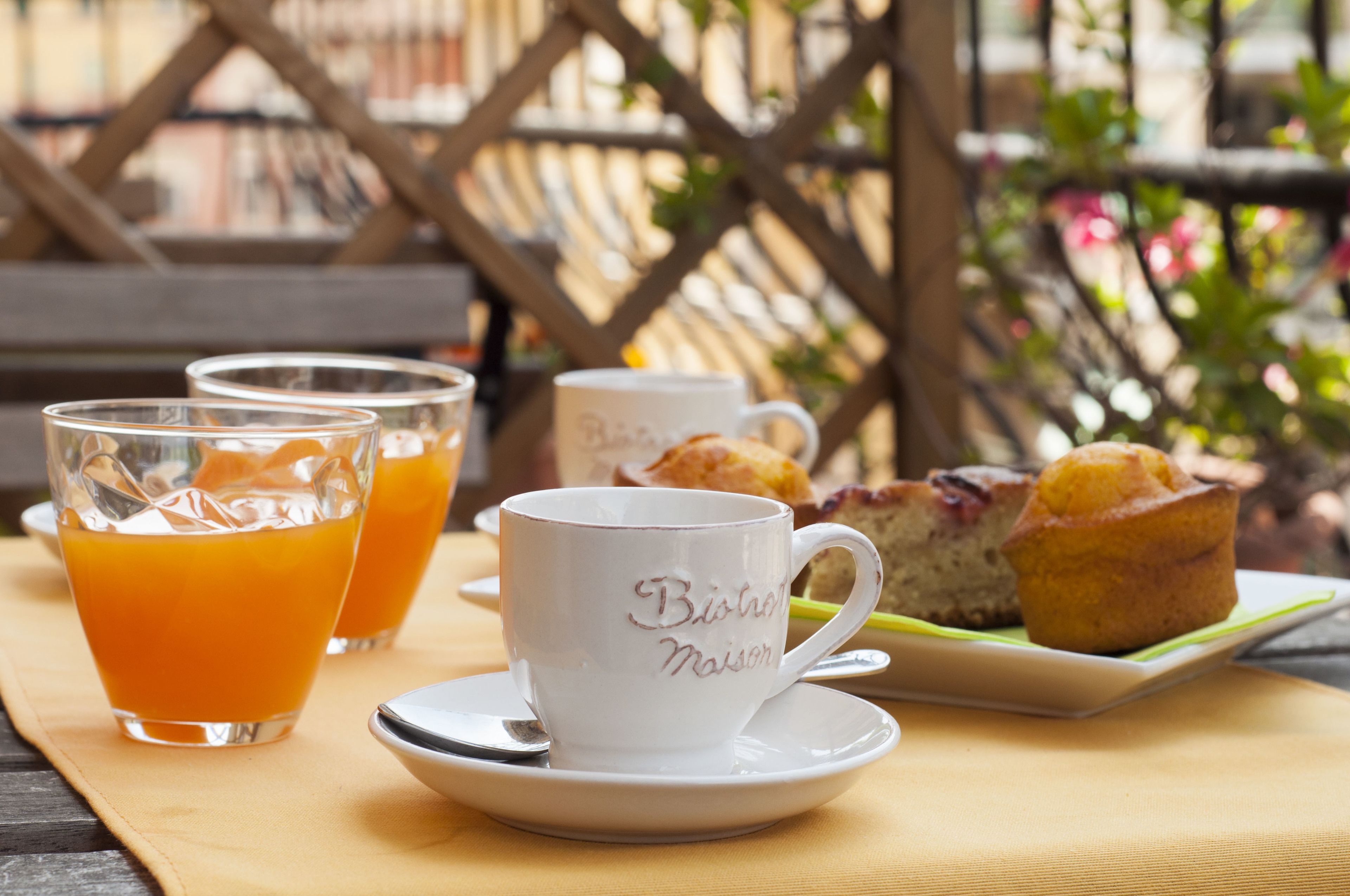 Ein Frühstückstisch mit einer weißen Tasse und Untertasse mit der Aufschrift "Bishop Maison", zwei Orangensaftgläsern sowie einem Teller mit Muffins und einem Gebäckstück.