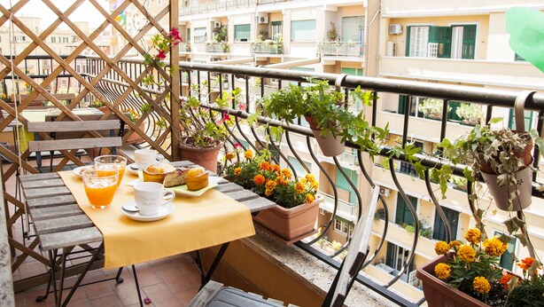Un balcon avec une table préparée pour le petit-déjeuner, des plantes en pot et une vue sur les bâtiments environnants.