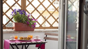 Un balcon avec une table préparée pour le petit-déjeuner, comprenant des muffins, du jus d’orange et une tasse. Une plante en pot aux fleurs violettes est posée sur le rebord de la fenêtre.