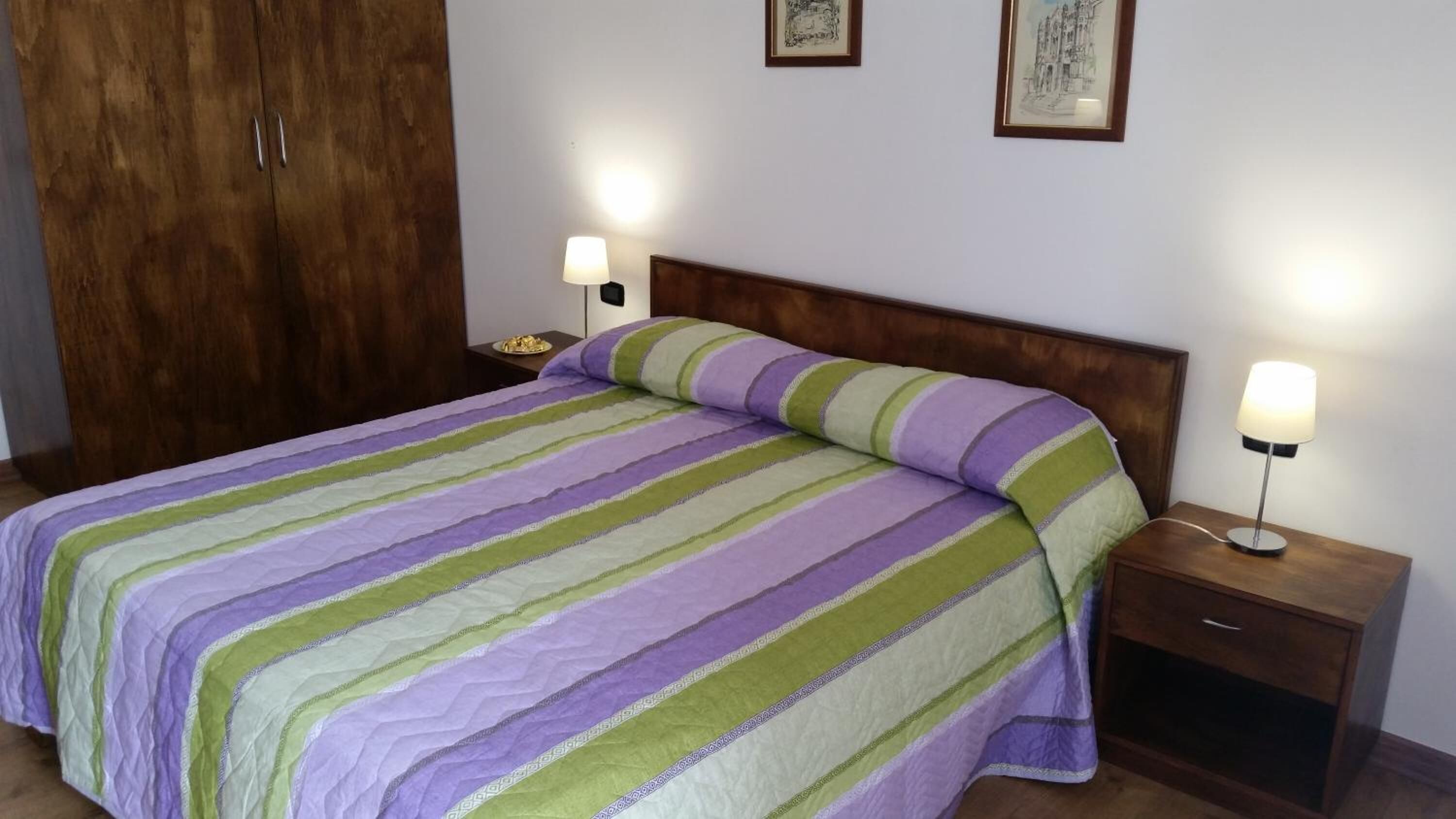 A neatly made bed with a striped bedspread, two bedside lamps, a wooden headboard, and a wardrobe.