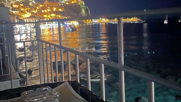 A night view from a balcony with a dining table, a bowl of fruit, and a view of a brightly lit coastal town. - B&B Il Borgo (Scilla)