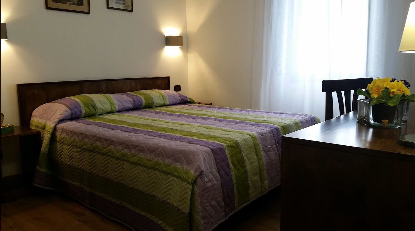 Una cama bien hecha con una colcha a rayas, una mesita de noche con una lámpara y una silla con un jarrón de flores.