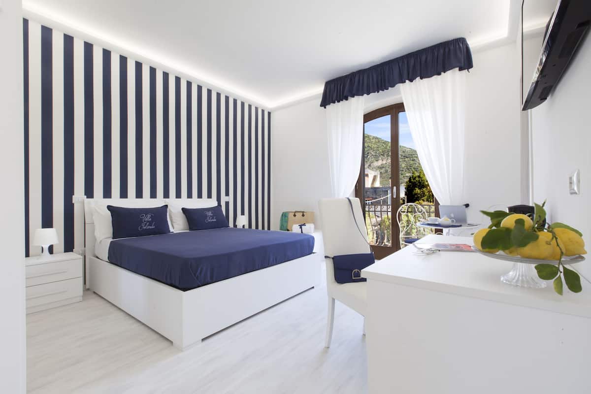 Un dormitorio con cama, mesita de noche y escritorio con silla. Hay una ventana con cortinas y vistas al exterior.