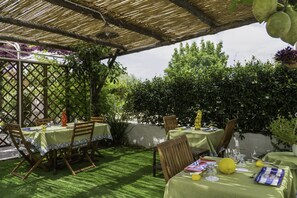 Un espace de restauration en plein air, avec des tables et des chaises disposées sous une pergola, entouré d’une végétation luxuriante et d’une clôture blanche.