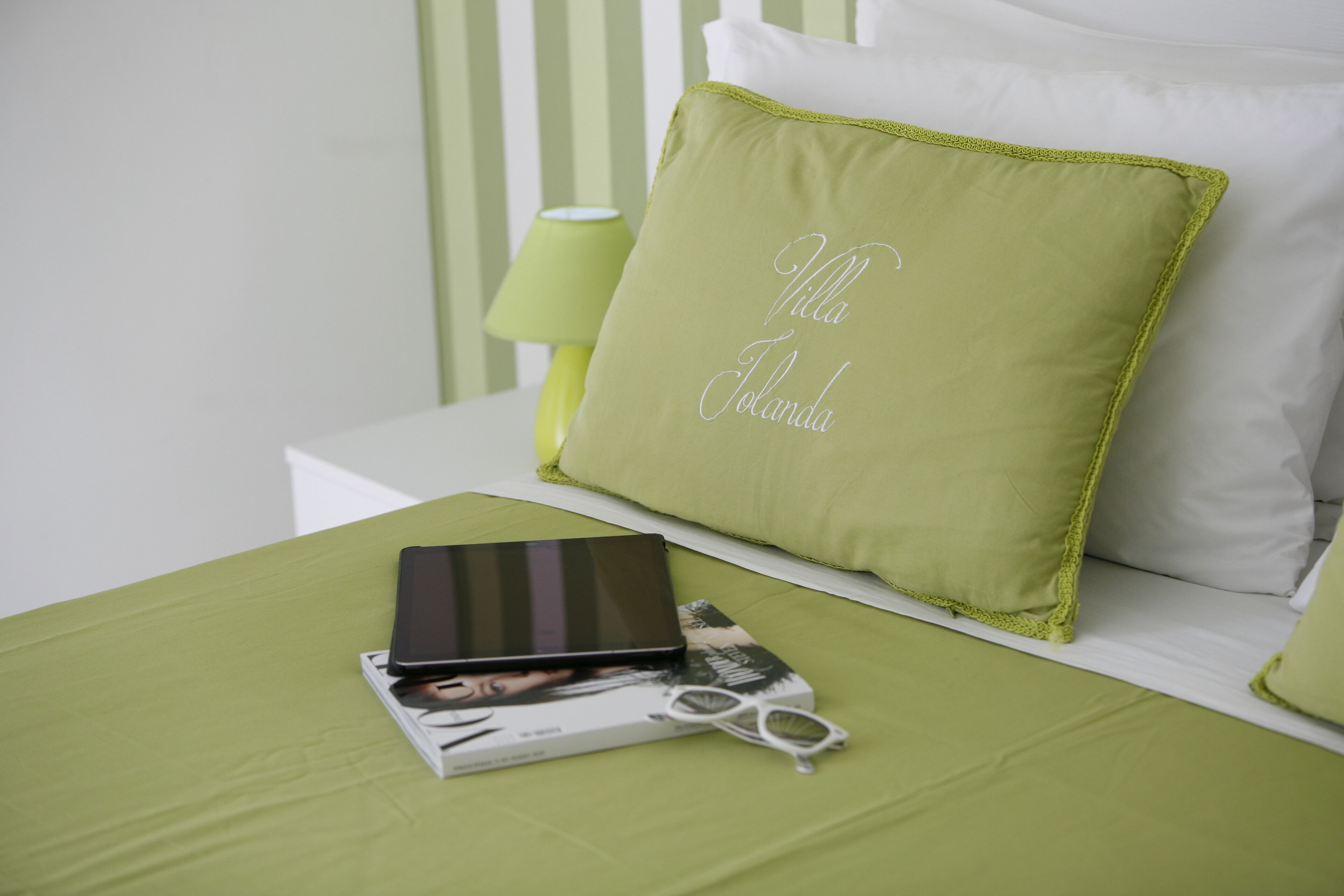Ein ordentlich gemachtes Bett mit einer grünen Tagesdecke, ein Kissen mit der Aufschrift "Villa Tolanda", ein Buch und eine Brille.