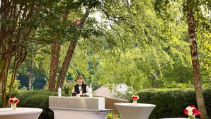 A garden setting with a reception desk and two round tables, surrounded by lush greenery and trees. - Sonesta ES Suites Gwinnett Place Atlanta (Duluth)