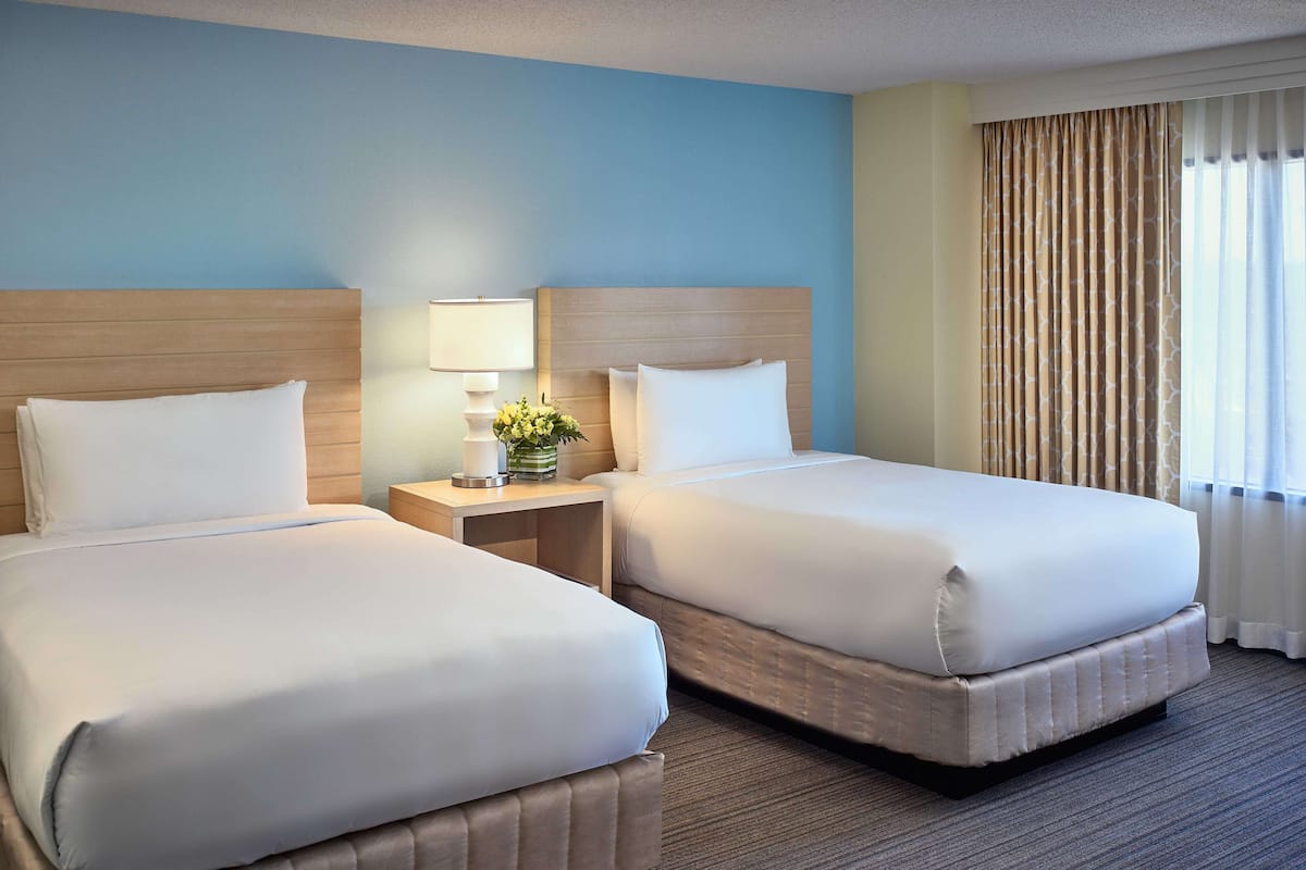 A hotel room with two beds, a nightstand with a lamp and flowers, and a window with curtains.
