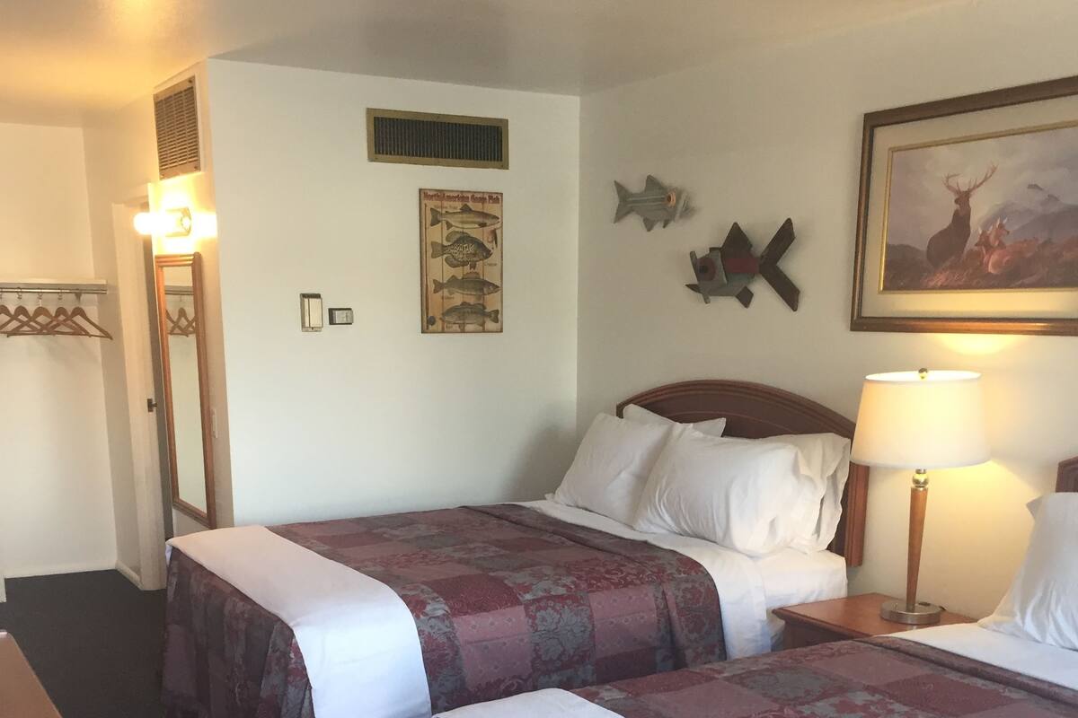 Une chambre d’hôtel avec deux lits, une tête de lit en bois, une table de chevet avec une lampe, une horloge murale, un tableau représentant un poisson et un porte-manteau.