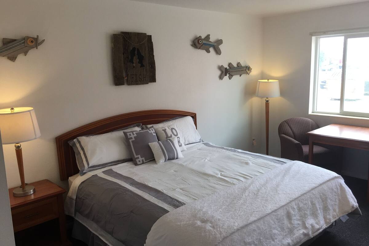 Un lit bien fait, avec une tête de lit en bois, une table de chevet avec une lampe et une chaise près d’une fenêtre.