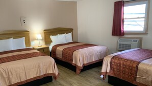 Habitación de hotel con tres camas, cabeceros de madera, una mesita de noche con lámpara, una ventana con cortinas y un equipo de aire acondicionado.