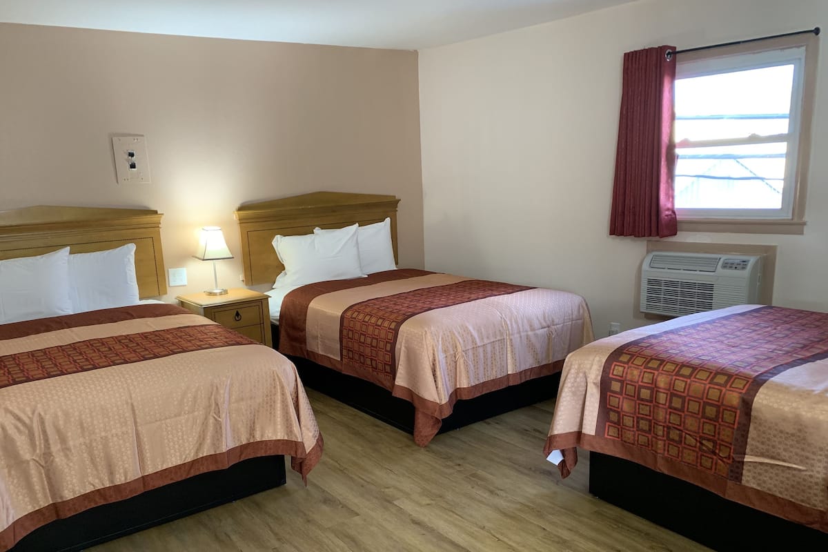 A hotel room with three beds, wooden headboards, a nightstand with a lamp, a window with curtains, and an air conditioning unit.