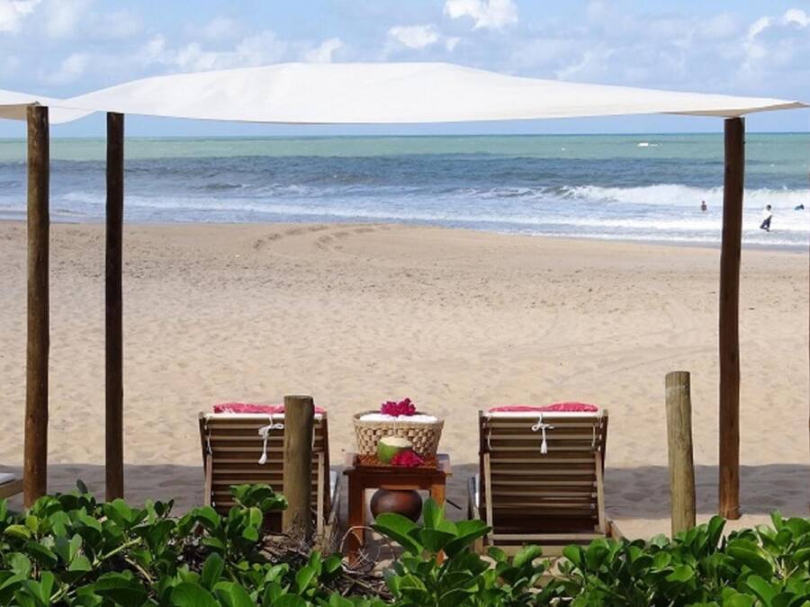 A beach with two wooden lounge chairs under a canopy, a beach chair with a basket and flowers, and a view of the ocean.