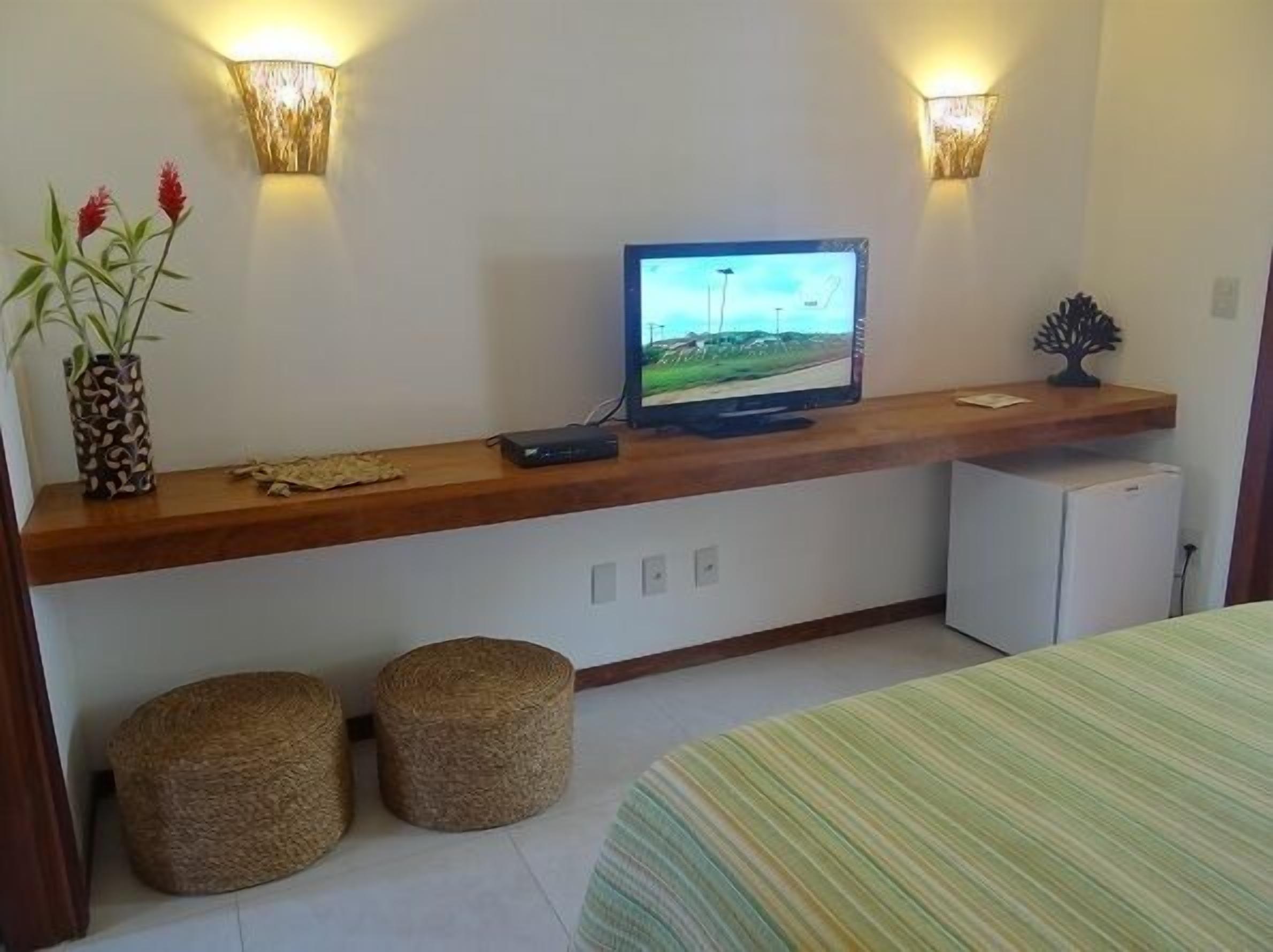 A room with a television on a wooden shelf, two wall-mounted lights, a small refrigerator, and two woven stools.