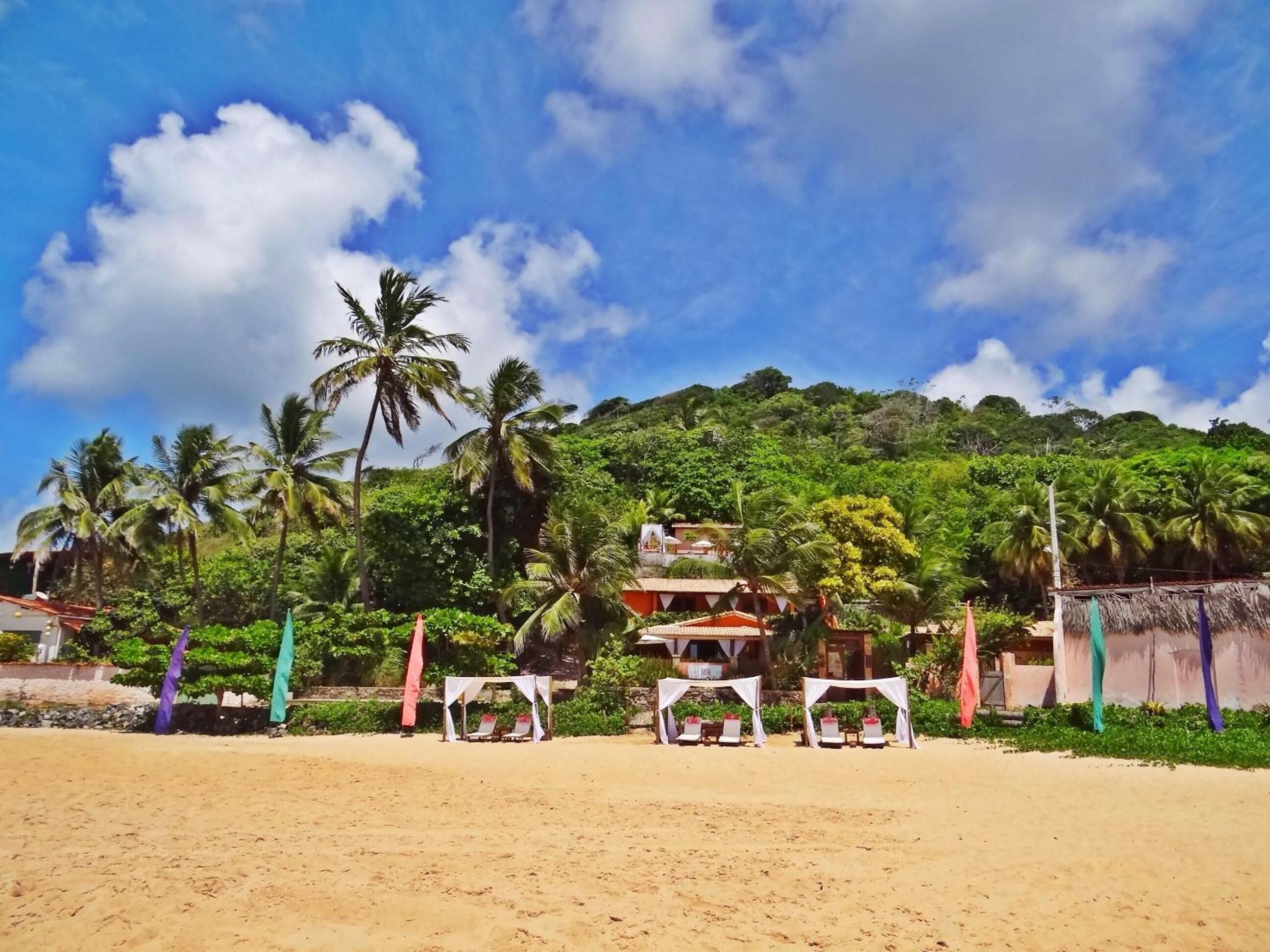 Uma praia com toldos brancos e cadeiras de praia, areal arenoso e um cenário de palmeiras e uma colina com edifícios.