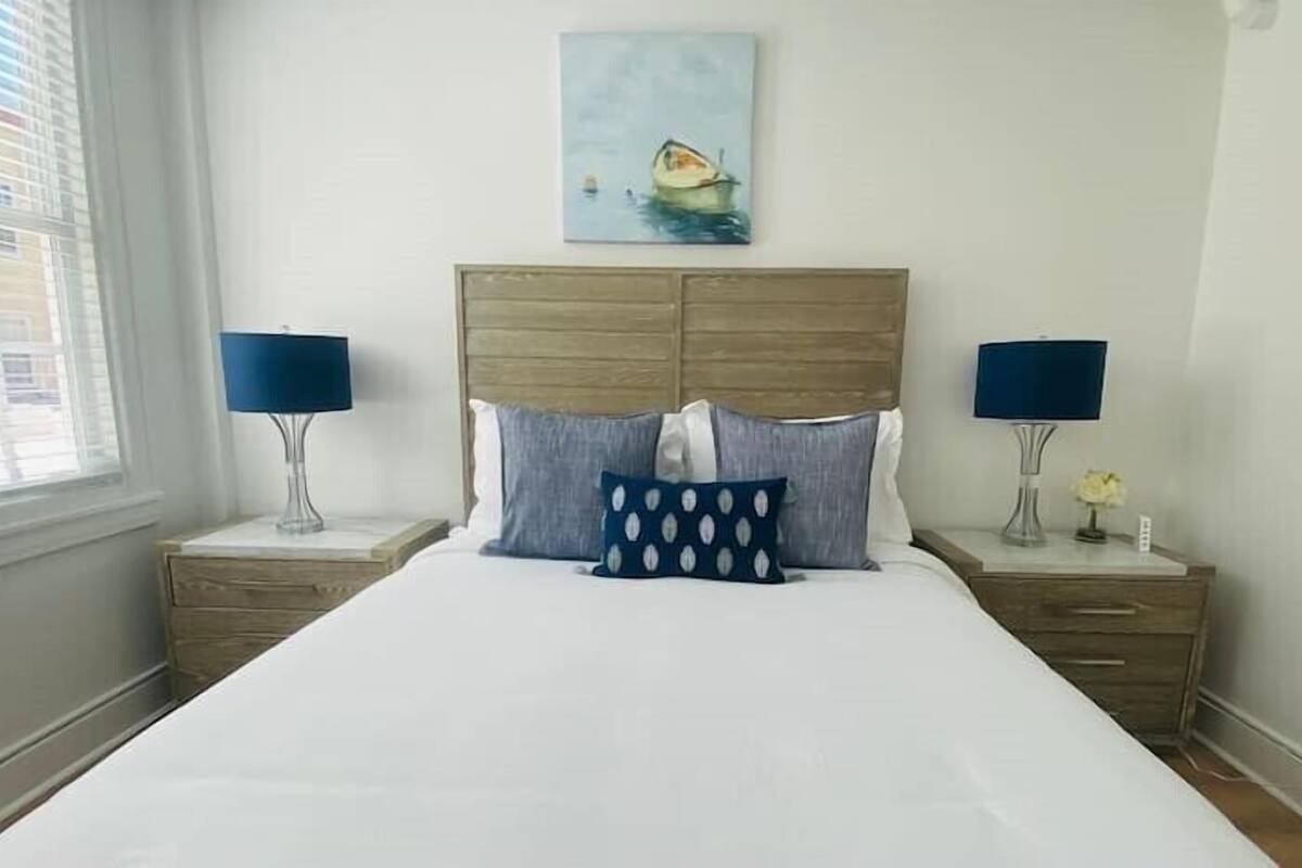 Ein ordentlich bezogenes Bett mit weißer Daunendecke und zwei Kissen, begleitet von Holz-Nachttischen mit Lampen und einer kleinen Vase. Über dem Bett hängt ein Bild von einem Boot.