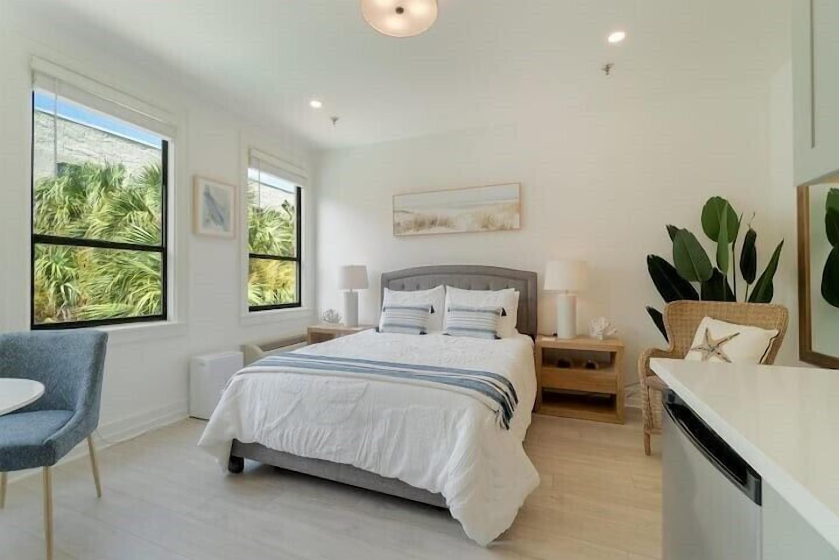 Une chambre moderne avec un grand lit, des tables de chevet, un fauteuil et une vue sur les palmiers par les fenêtres.