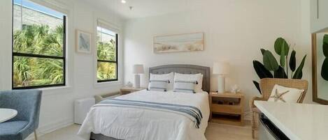 Une chambre moderne avec un grand lit, des tables de chevet, un fauteuil et une vue sur les palmiers par les fenêtres.