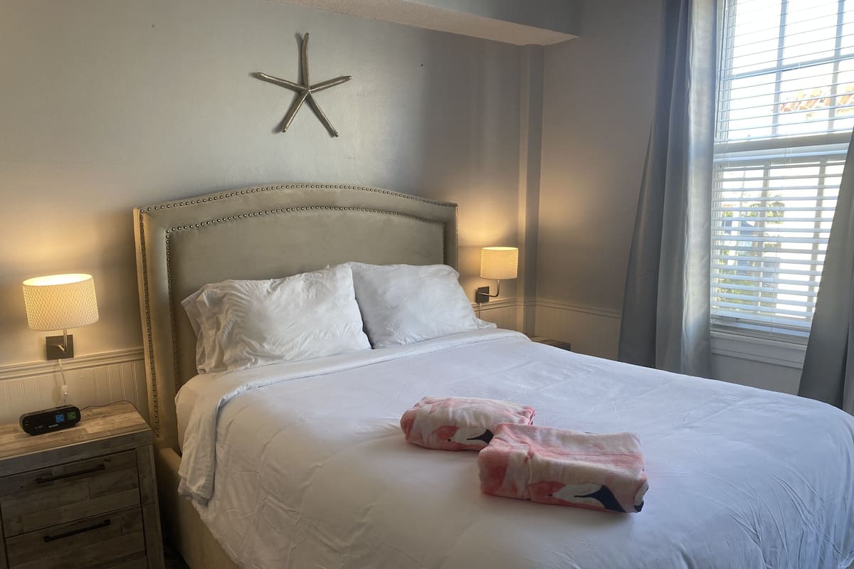 Ein ordentlich bezogenes Bett mit weißen Laken und pinkfarbenen Handtüchern, eine Meerstern-Dekoration an der Wand und zwei Nachttischlampen.