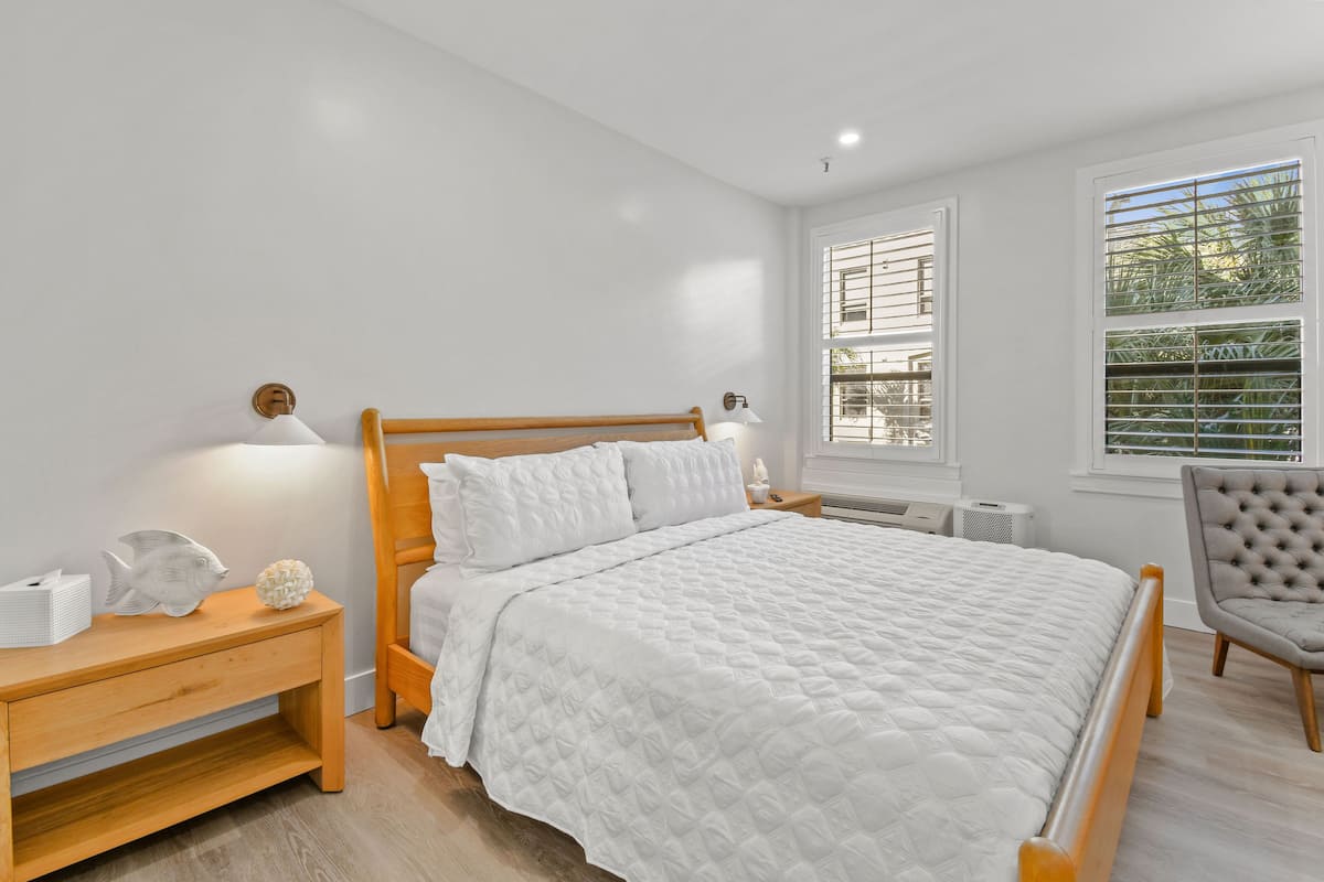 Ein Schlafzimmer mit einem hölzernen Bett, weißer Bettwäsche, einem Nachttisch mit Lampe, einem Sessel und einem Fenster mit Jalousien.