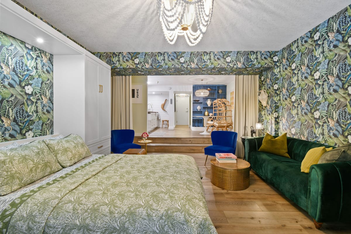 Ein Schlafzimmer mit einem Bett, einem grünen Sofa und einem Sessel. Eine Tür führt zu einem Badezimmer und einem Flur.