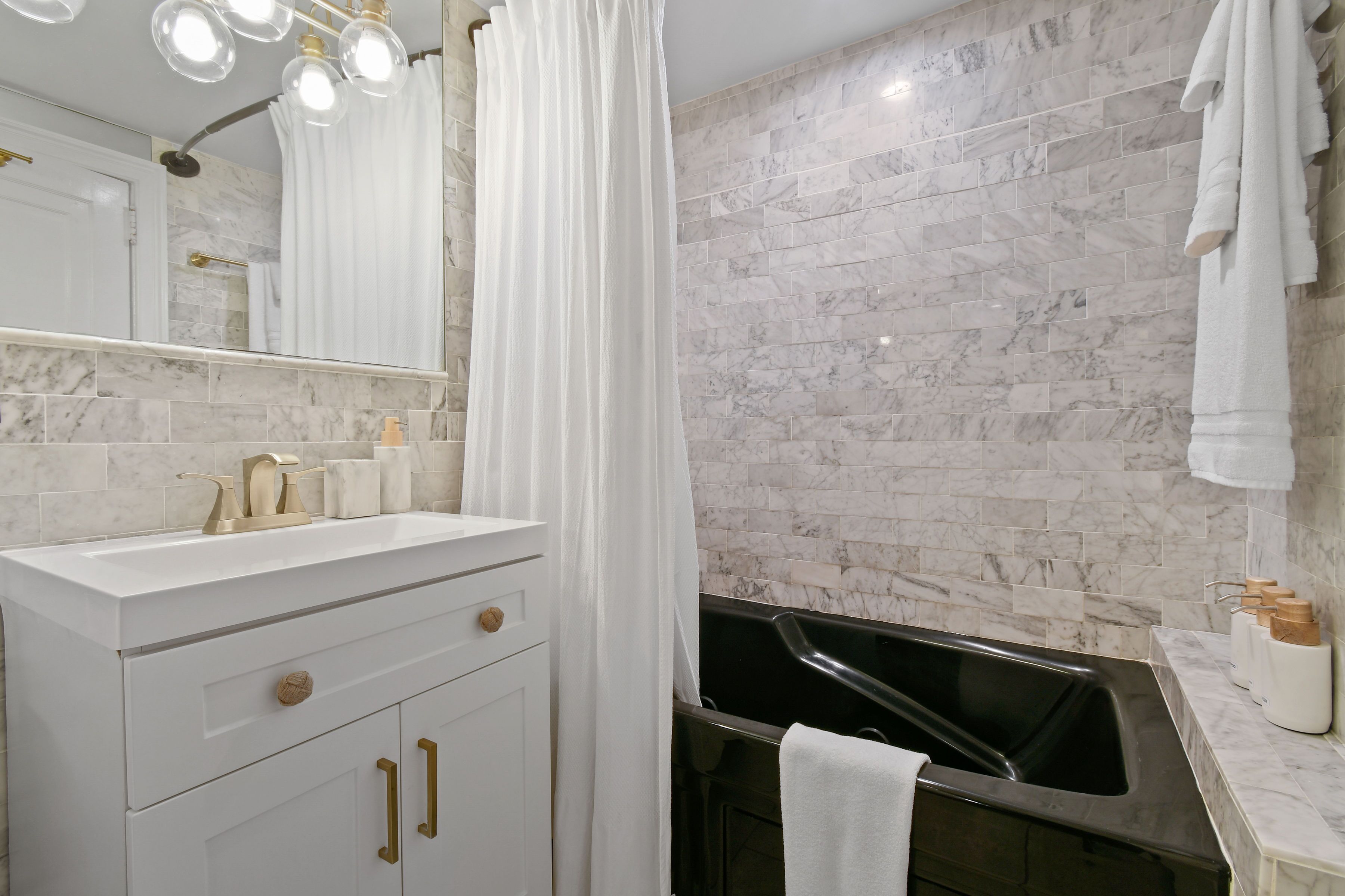 A modern bathroom with a white vanity, a large mirror, a bathtub, and marble walls.