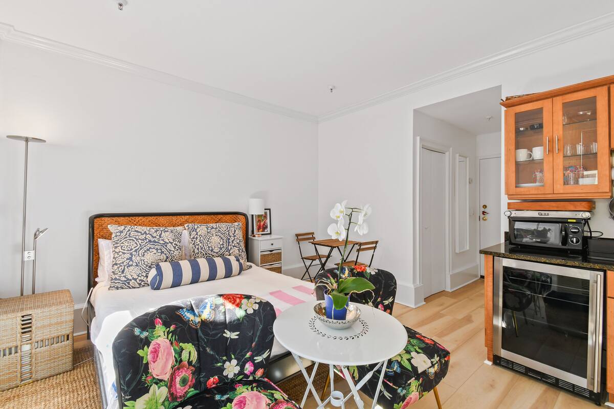 Ein Schlafzimmer mit Bett, Nachttisch, Sessel, einem kleinen Tisch mit Pflanze sowie einer Küchenzeile mit Mikrowelle und Schränken.
