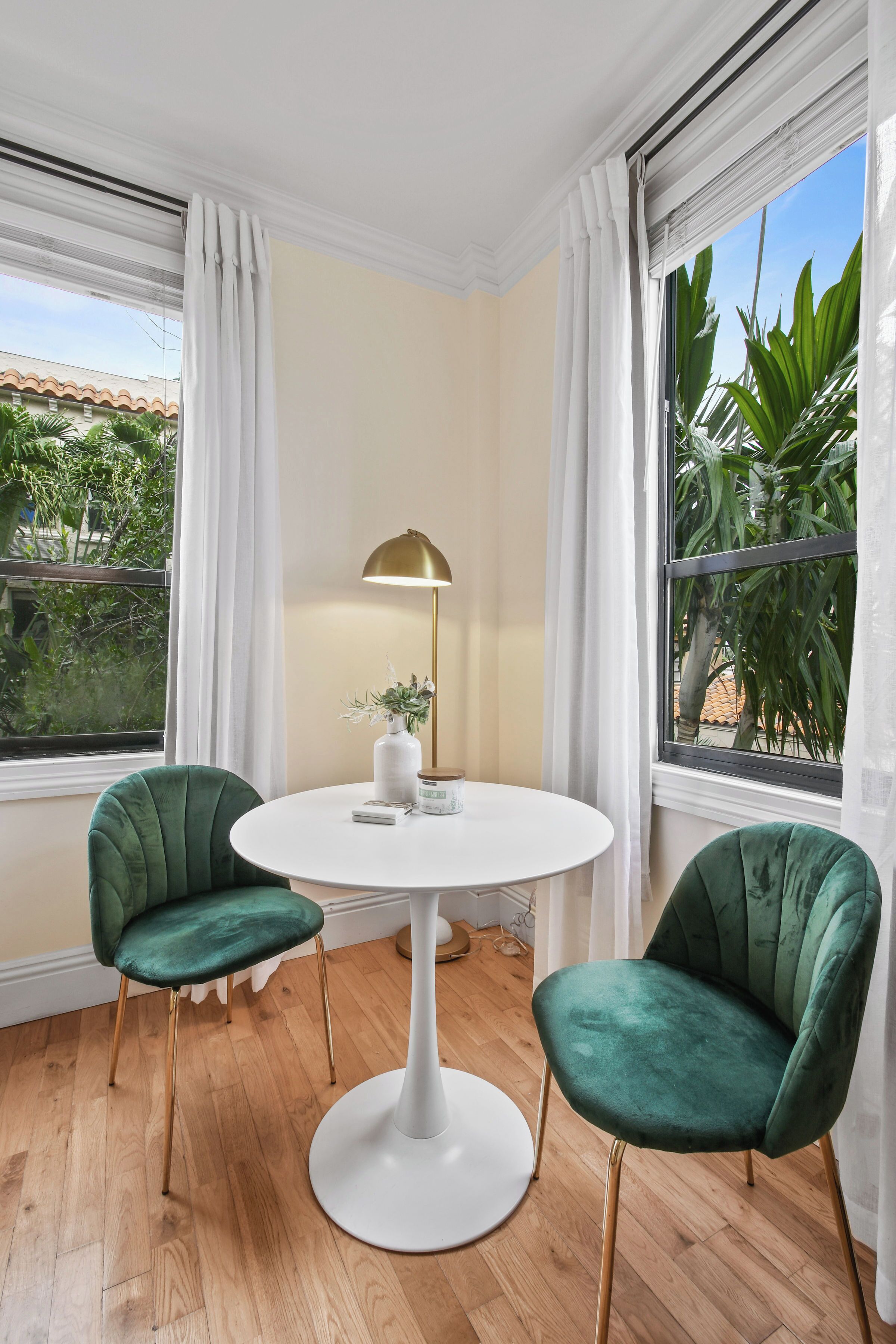 A room with a round white table, two green chairs, a gold floor lamp, and a vase with flowers.