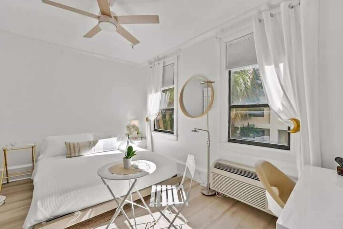 Ein heller, weißer Raum mit einem Bett, einem runden Tisch und einem Stuhl. Es gibt einen Deckenventilator, ein Fenster mit Vorhängen und einen wandmontierten Spiegel.
