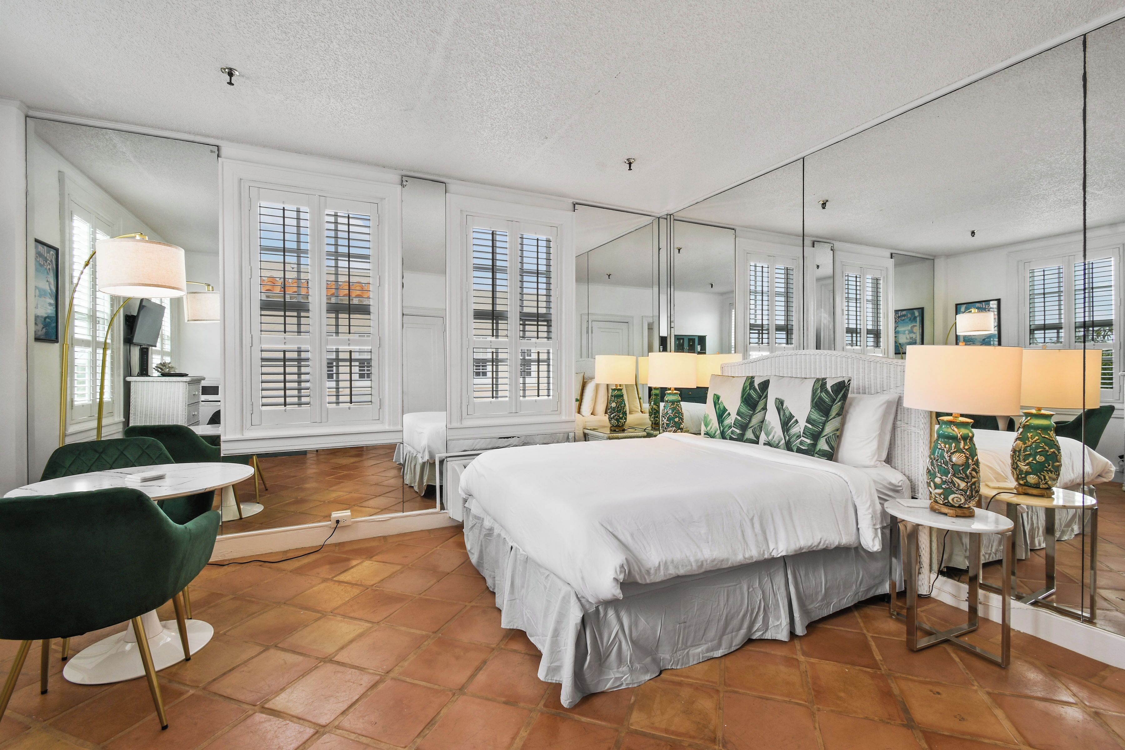 A hotel room with a large bed, two bedside tables, a green armchair, a small round table, and a large mirror.