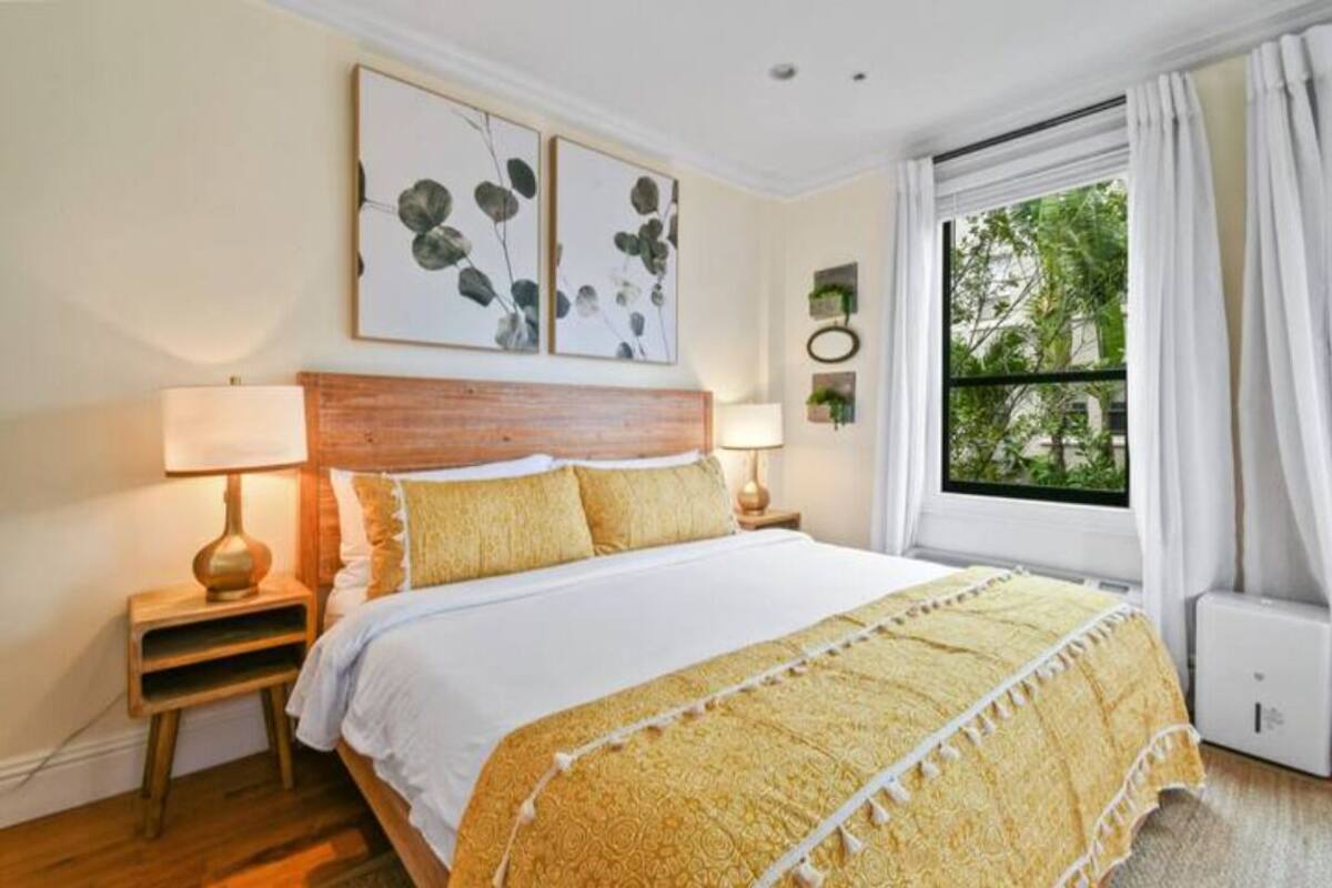 Ein Schlafzimmer mit einem Holz-Kopfteil, einem großen Bett mit gelbem Musterüberwurf, zwei Nachttischlampen und einem Fenster mit weißen Vorhängen.