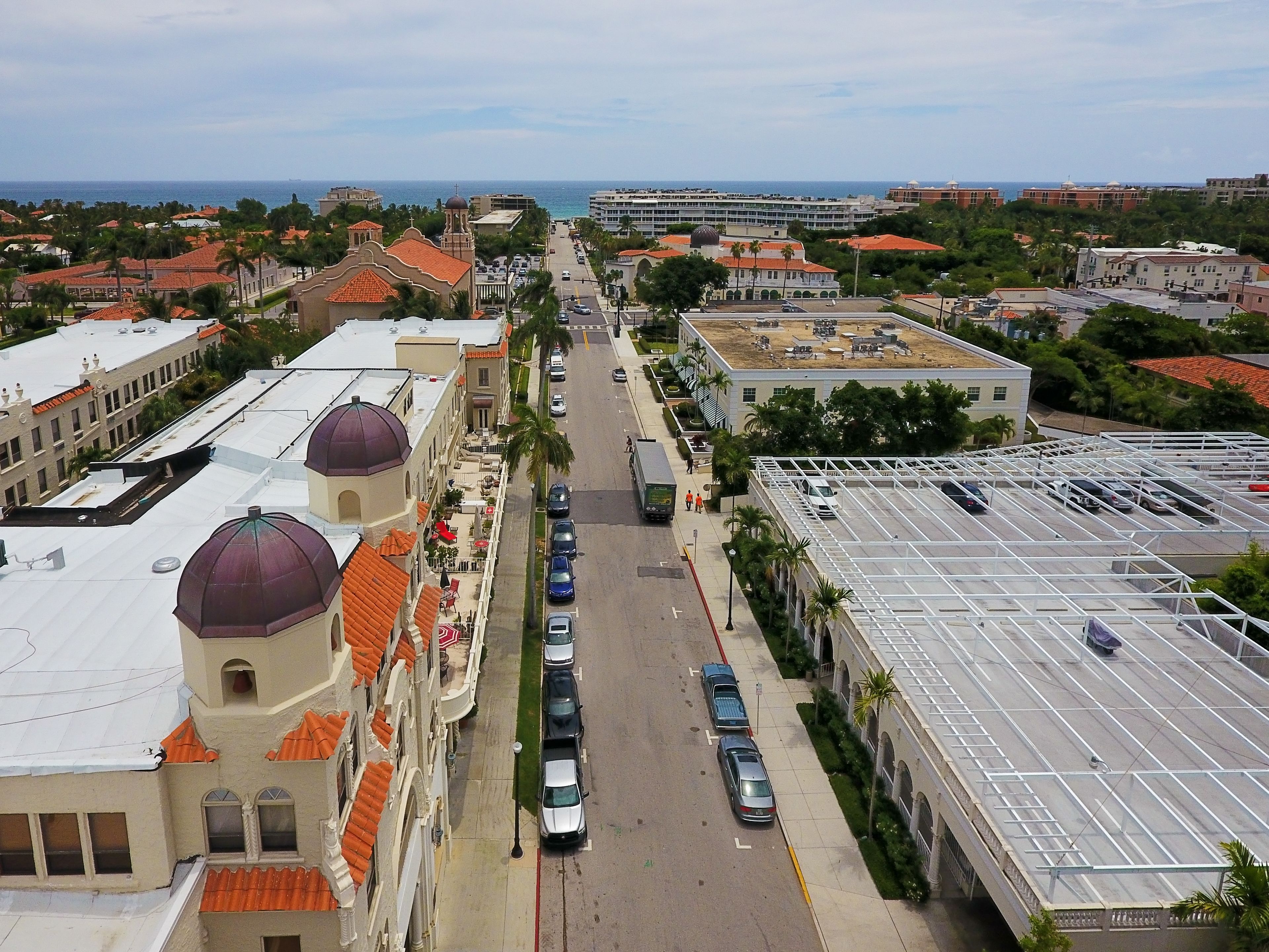 Vista aérea de una ciudad costera con edificios de techos rojos y estructuras abovedadas, una calle amplia y automóviles estacionados.