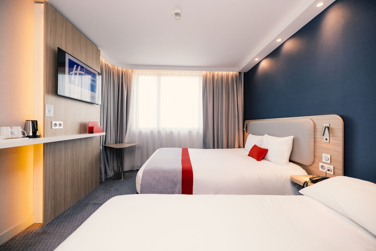 Ein Hotelzimmer mit zwei Betten, einem Fernseher, einem Schreibtisch mit Kaffeemaschine und einem Fenster mit Vorhängen.