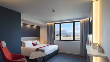 Een hotelkamer met een bed, een rode stoel, een bureau met een kopje en een groot raam met uitzicht op de stad.