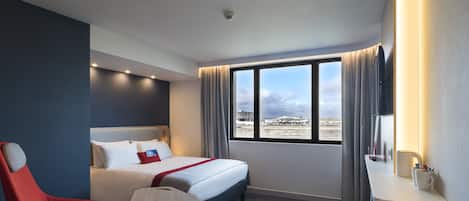 A hotel room with a bed, a red chair, a desk with a cup, and a large window with a view of the city.