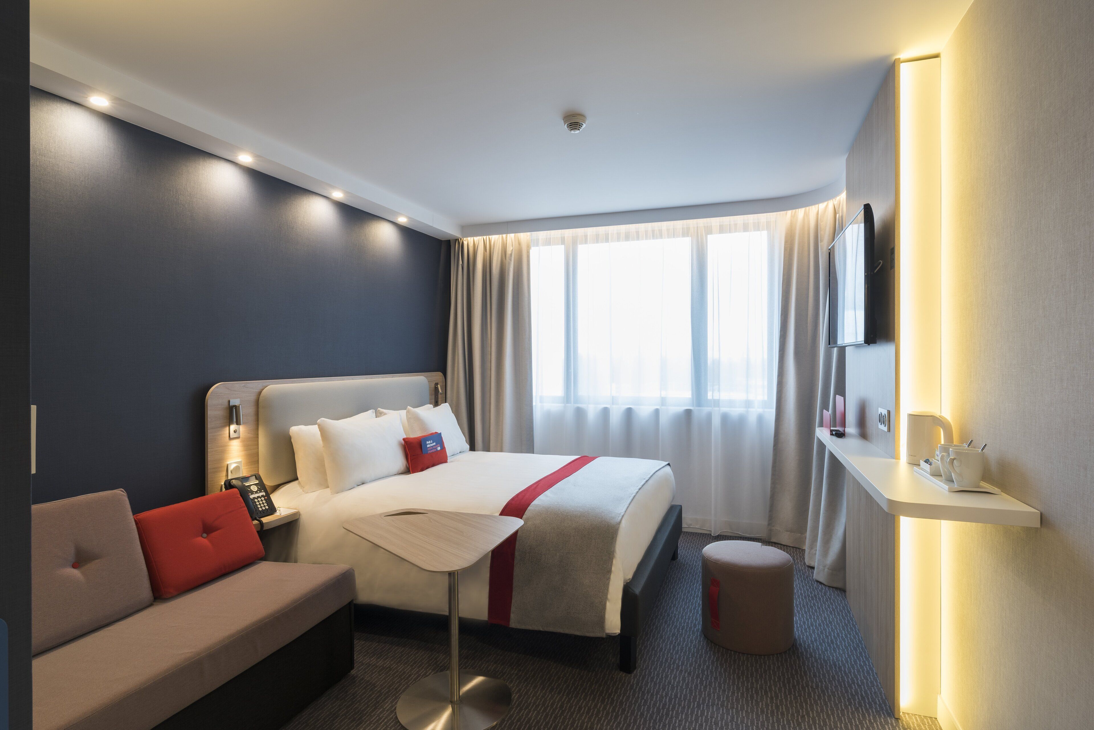 A hotel room with a bed, a sofa, a bedside table with a lamp, a TV, and a small table with a cup and spoon.