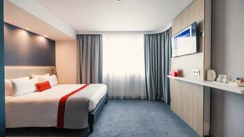 A hotel room with a bed, a TV, a desk, and a window with curtains.