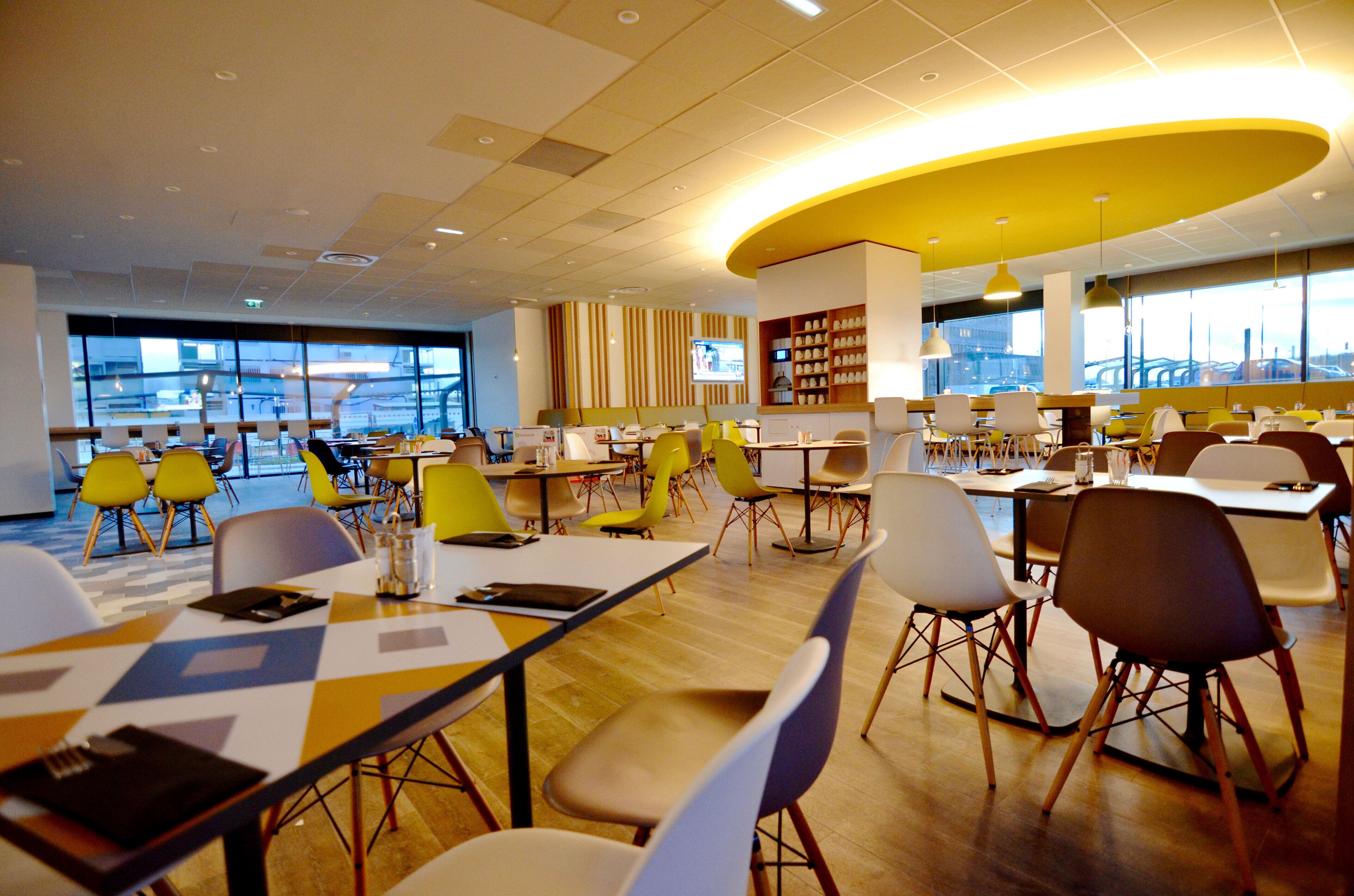 A modern dining area with multiple tables and chairs, large windows offering views of the city, and a curved yellow ceiling feature.
