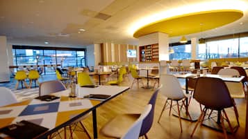 A modern dining area with multiple tables and chairs, large windows offering views of the city, and a curved yellow ceiling feature.
