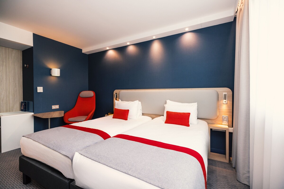 Ein Hotelzimmer mit zwei Betten, einem roten Sessel, einem Nachttisch und einer Wandlampe.