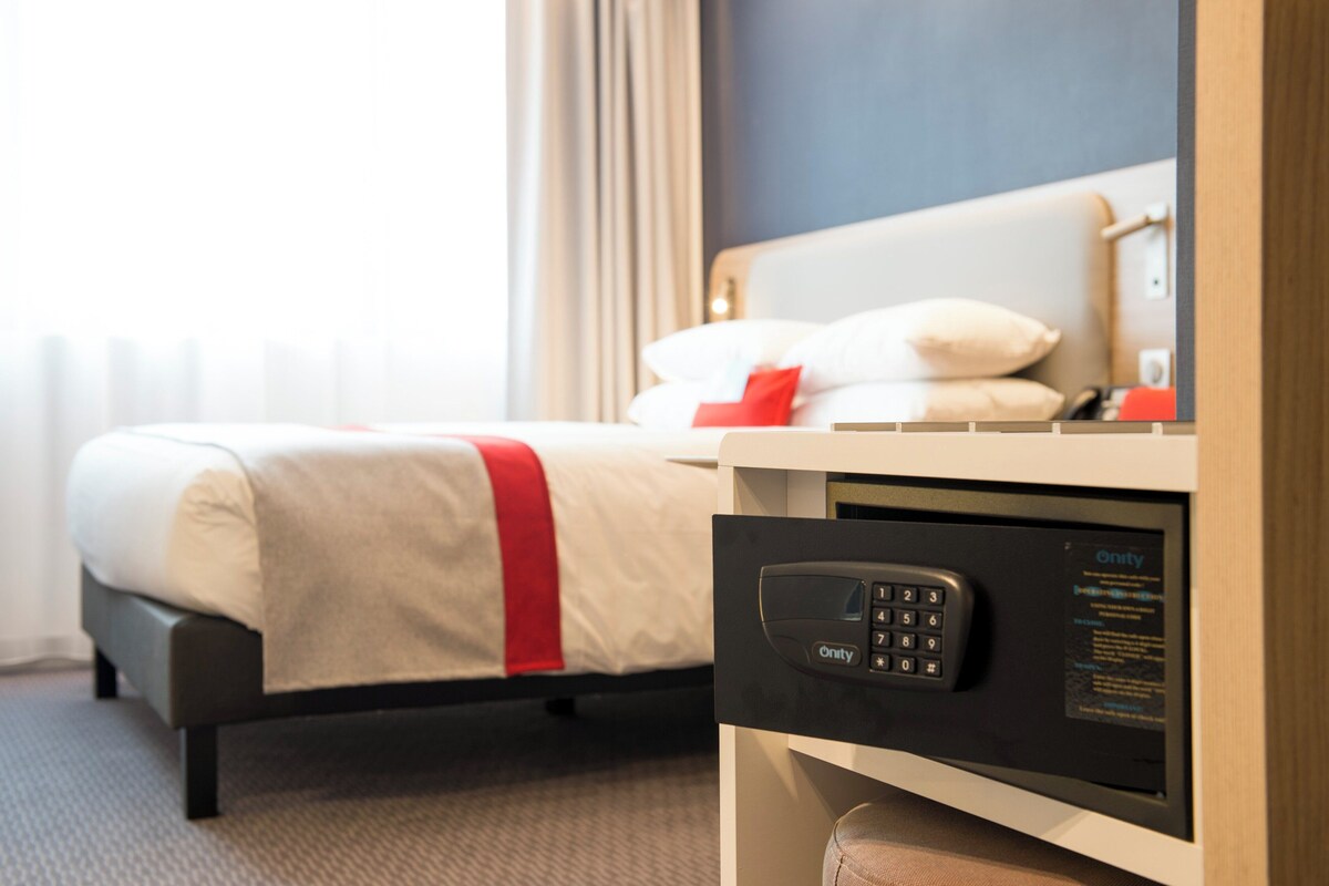 A hotel room with a bed, a bedside table with a control panel, and a window with curtains.