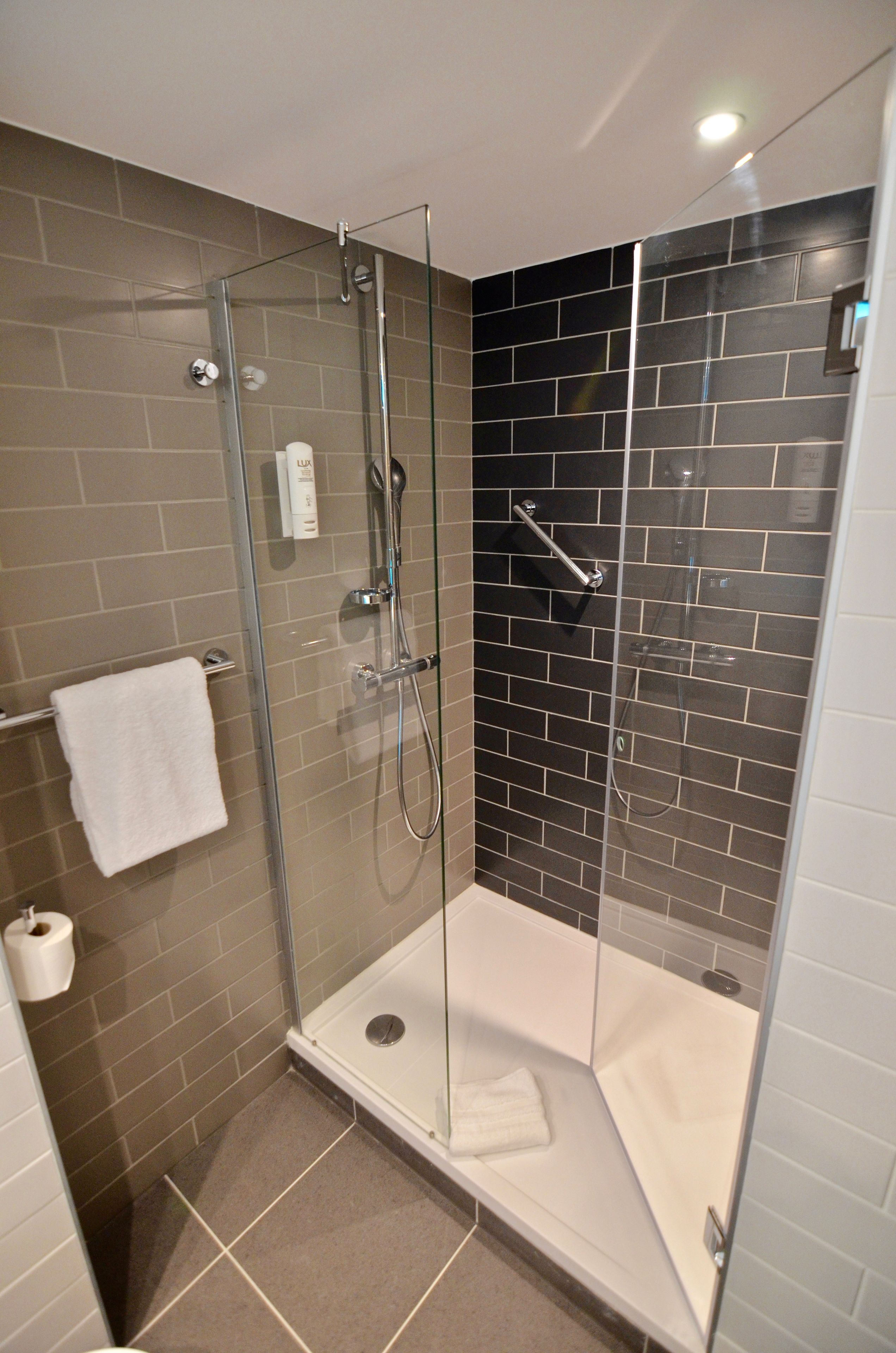 Een douche met glazen wand, een wit bad, wandtegels in beige en zwart, en een witte handdoek op de vloer.