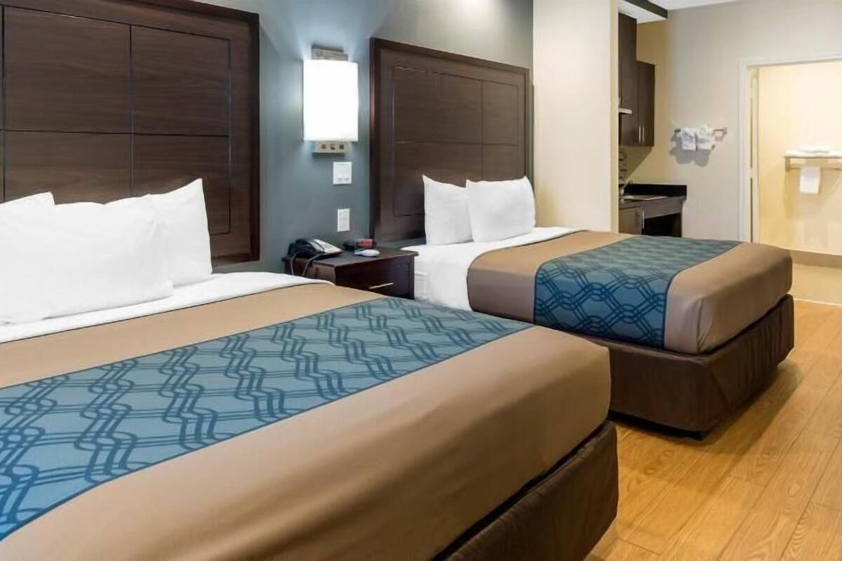 A hotel room with two beds, wooden headboards, a bedside table with a phone, and a small desk with a lamp.