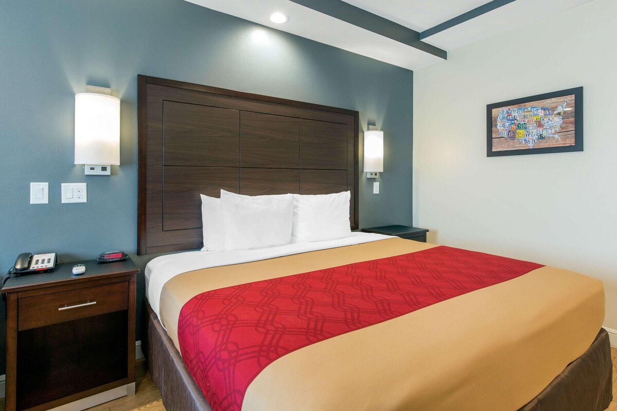 A hotel room with a large bed, a nightstand with a phone and a clock, and a wall-mounted light fixture.