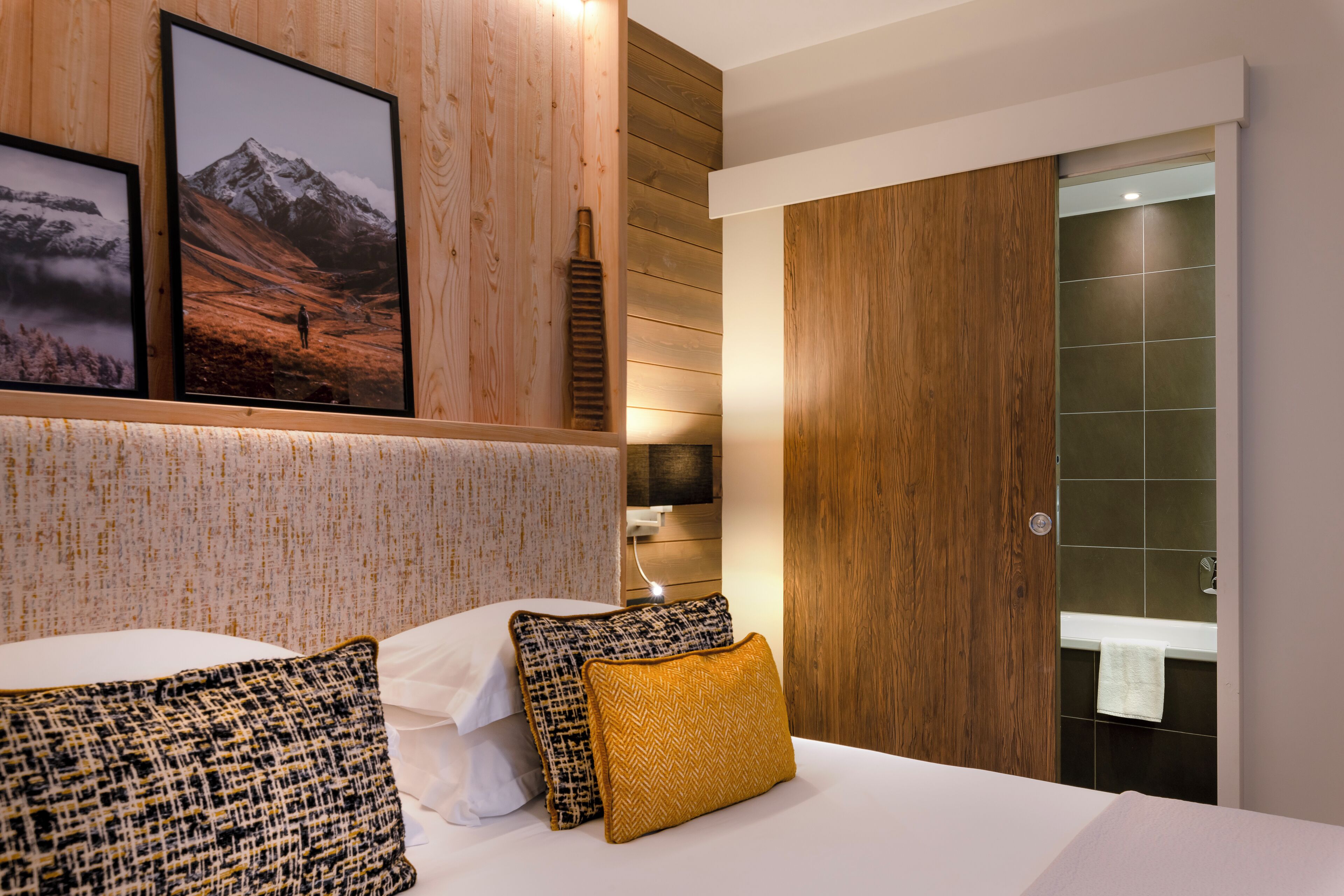 A hotel room with a wooden headboard, a bed with white linens, two patterned pillows, and a framed photograph of a mountain.