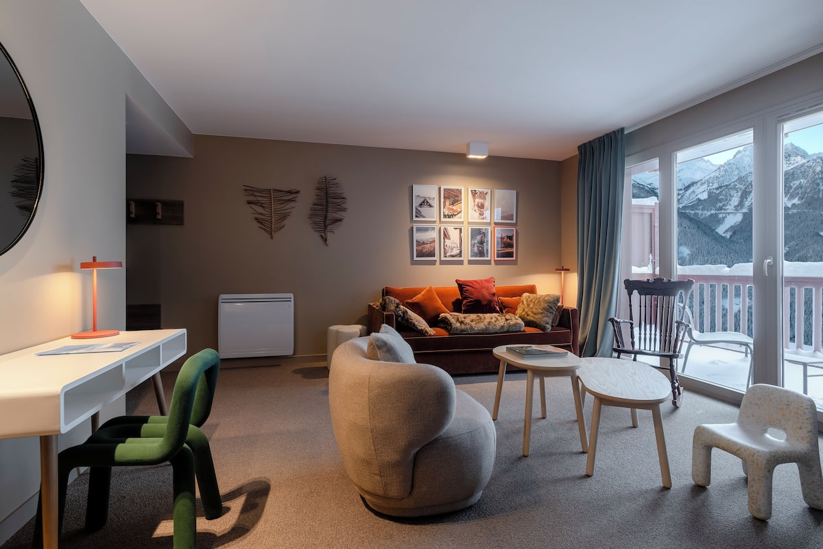 Ein modernes Wohnzimmer mit einer Couch, Sesseln, einem Schreibtisch und Blick auf einen verschneiten Berg.